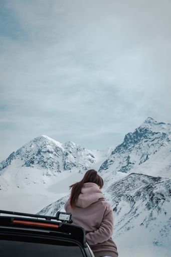 A female leaning on a black vehicle in front of amazing snowy and rocky mountains and cloudy skies