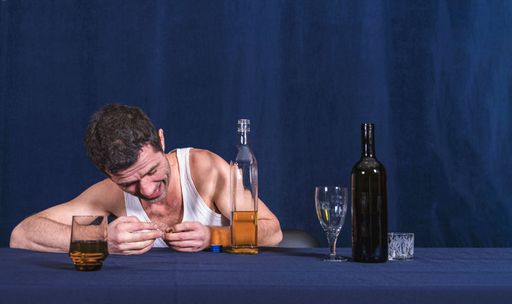 Lonely drunk man in homely white T-shirt sits at table and rips off wedding ring from his finger. In front of him are bottle of alcohol and glasses. A