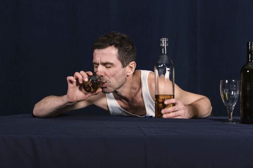 Lonely drunk man in homemade white T-shirt lies on table and drinks alcohol from glass. Other hand holds bottle of vodka. Alcoholism as disease.