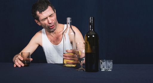Lonely and drunk man in white home shirt sits at table. In his hands holds glass and bottle of vodka. Drunken look is directed at bottle of alcohol.