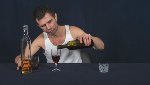 Lonely and drunk man in white home shirt sits at table He holds bottle of alcohol in his hand and pours wine from it into glass Alcoholism as disease