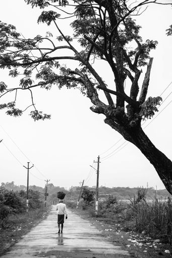 A vertical grayscale shot of a lonely man carrying a bucket on his head while walking on the road