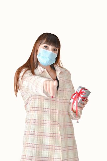 Young woman wearing a surgical mask holding a silver gift box and pointing at the camera on an out of focus background. Gifts and safety concept.