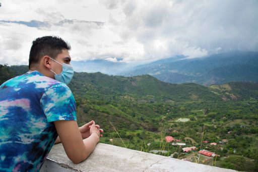 A back view of a Hispanic young man in a medical face mask leaning to the stone wall and enjoying a beautiful nature scene