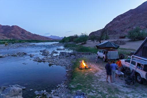 Two camping trucks parked near the lake and folding chairs with a campfire in front of it