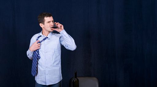 White man in business shirt stands against dark background and drinks red wine from glass with large gulps. He rips his tie off neck with hand. Stress