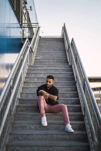 A young hot man wearing a black oversize shirt and pink pants sitting on a staircase