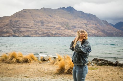 A blonde female with a denim jacket standing near the wavy sea surrounded by high rocky mountains