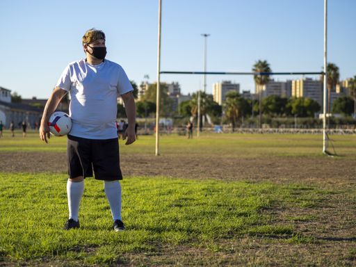 A young athlete with a white shirt and black mask holding a football ball and posing for the camera