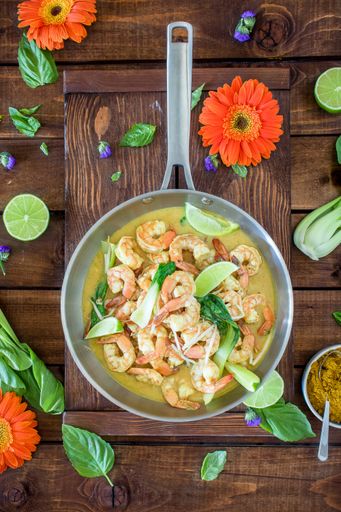 Curry shrimp soup served in a silver pot on a brown table in a composition with flowers, leafs and fruits