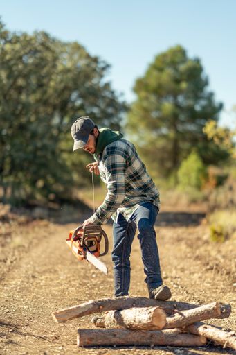 A vertical shot of a young Caucasian man cutting a tree trunk with a chainsaw