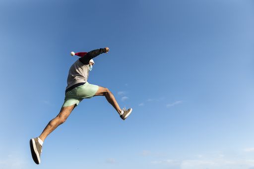 A low angle shot of a young Hispanic man wearing a Santa hat and a face mask, jumping under the blue sky, parkour during Christmas, COVID-19