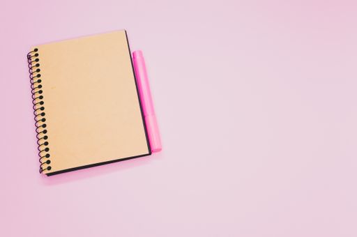 A high angle shot of a notebook and a pink marker on a pink surface