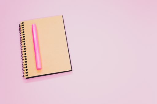 A high angle shot of a notebook and a pink marker on a pink surface