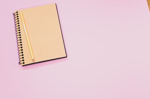 A high angle shot of a notebook and a pencil on a pink surface