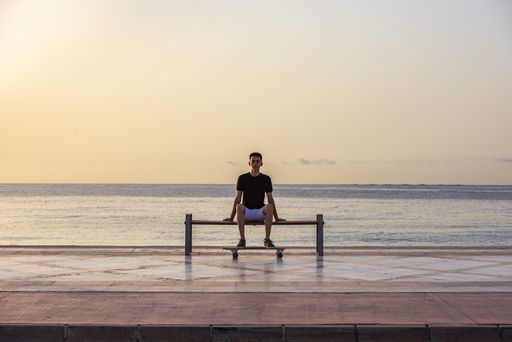 A selective focus shot of a handsome young teenager with his longboard sitting on a bench near an ocean
