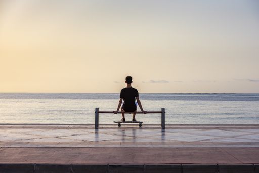 A selective focus shot of a handsome young teenager with his longboard sitting on a bench near an ocean