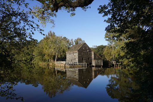 Historic Yates Mill reflecting in the millpond under a blue sky framed with tree leaves.  Located in Raleigh, North Carolina.