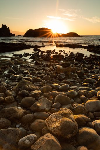 A vertical shot of a majestic golden sunset with sun rays over a rocky coastline and wavy seawater