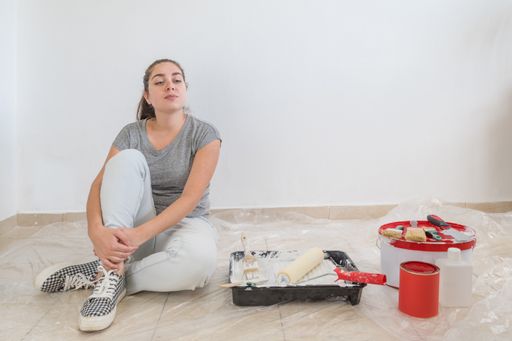A tired young woman sitting on the floor with a paintbrush and bucket while doing the repair at home