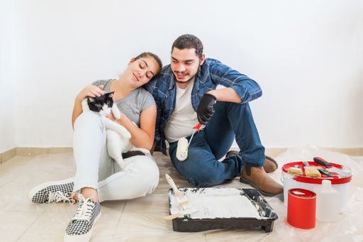 A tired young couple with a cat sitting on the floor with a paintbrush and bucket while doing the repair at home