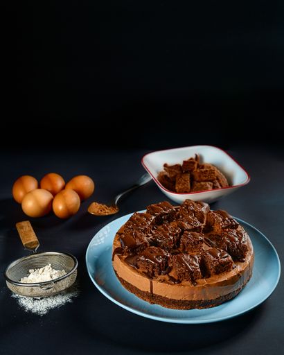 A vertical shot of a delicious chocolate cheesecake with small pieces of brownies on top, placed on a dark surface next to the ingredients