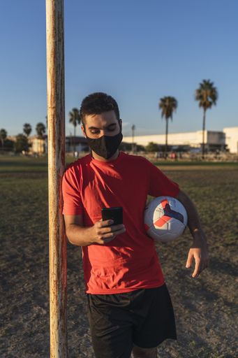 A soccer player in a mask scrolling on the phone during the break, the concept of a healthy lifestyle in the "new normal" conditions