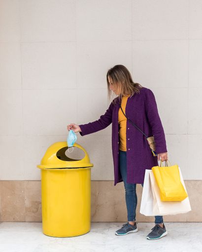 A vertical shot of a young woman carrying paper bags throwing sanitary mask in the trash can