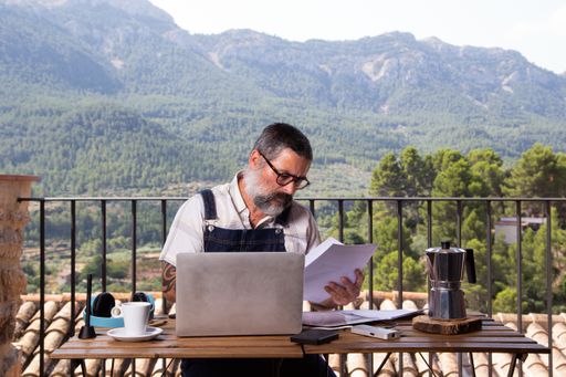 A selective focus shot of a man working on a laptop on the balcony of his house with nature in the background