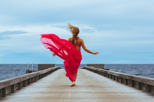 A soft breeze hitting the woman walking on a wooden pier wearing an elegant pink dress