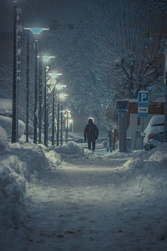 A vertical back view of a pedestrian walking on the sidewalk during snowstorm in the evening