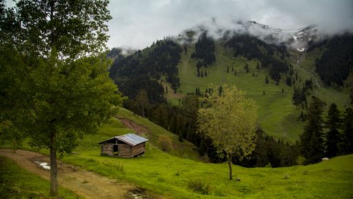 A wooden barn on the hill surrounded by greenery and mountains covered in forests and fog