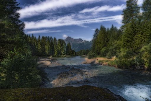 A river in a forest covered in greenery surrounded by hills under a blue cloudy sky and sunlight