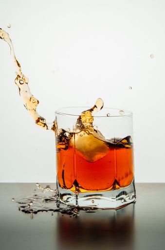 A vertical shot of a lowball glass of splashing whiskey with an ice piece on a reflective surface with spilled liquid droplets