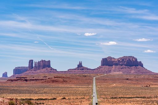 An aerial view of a long road in a desert leading towards ancient rock formations under a sunny sky