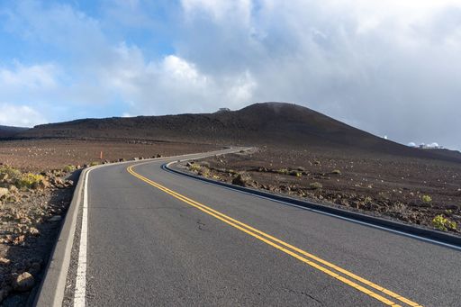 An empty curving road in the wilderness surrounded by dry fields and low hills under a bright cloudy sky