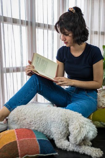 A Hispanic woman reading a book while sitting on colorful cushions around her dog