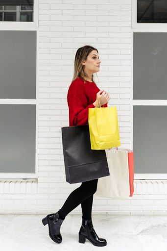 An adult Caucasian woman wearing a red sweater walking through a mall with colorful shopping bags
