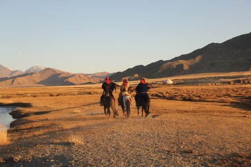 A landscape of a deserted valley surrounded by rocky mountains and a river with three horseriders