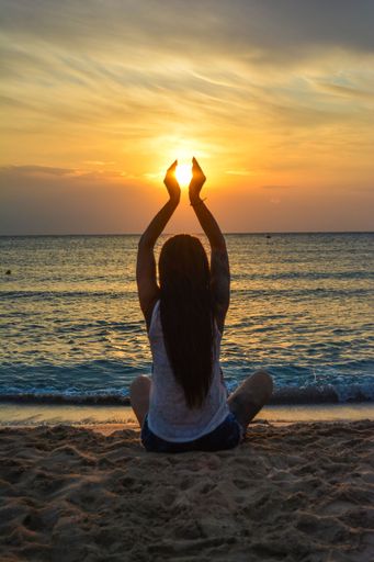 A relaxed young woman sits on a sun-drenched beach with her legs crossed, doing yoga