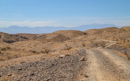 A rural dirt road crosses hills heading toward a mountain range in the arid California, desert.
