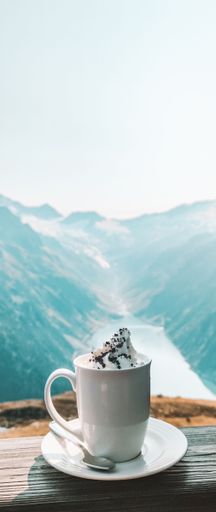 A vertical shot of a cup of coffee with cream on a window with the rocky mountains in the background