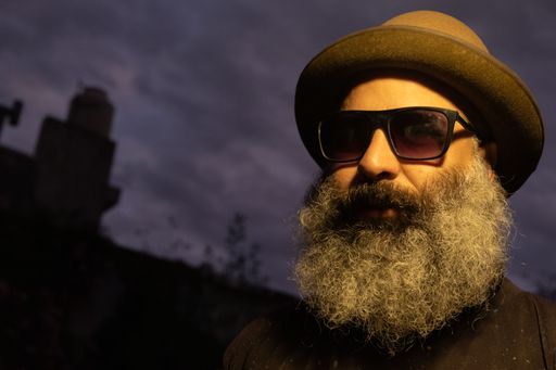 A caucasian beard man with a black hat and sunglasses against blurred dark sky in the evening
