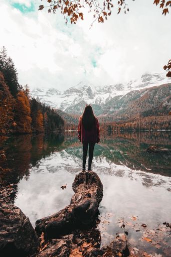 A vertical shot of a female standing on a big stone and looking at beautiful lake and colorful trees