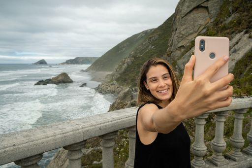 a Young woman cheerfully taking a selfie with a mountainous coast in northern Spain.