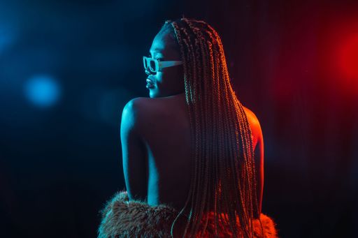 A portrait of an African young woman with braids wearing a coat posing under red and blue neon lights