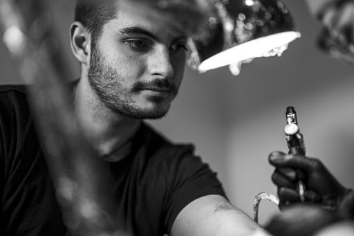 A grayscale shot of a young man in a tattoo salon tattooing his arm