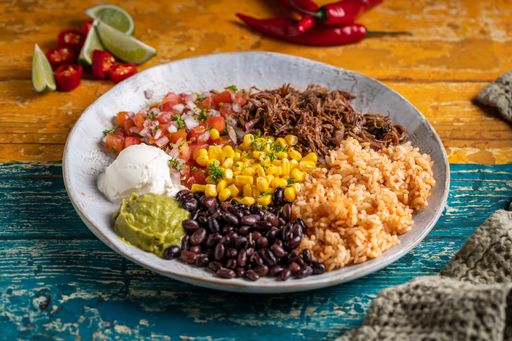 A Mexican dish of a barbacoa burrito bowl, meat rice black beans  sour cream guacamole and chopped vegetables
