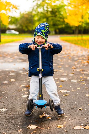 Young happy boy with a three wheeled scooter standing on a footpath at a park in the autumn season.