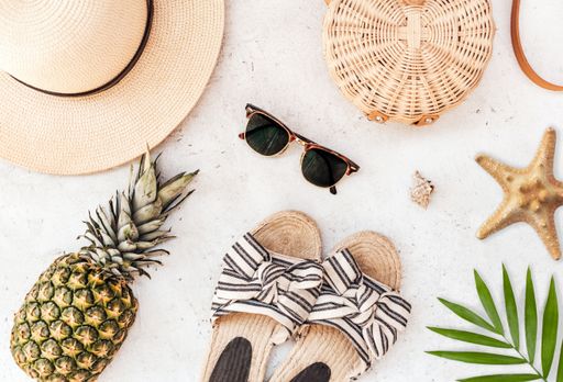 A flat lay shot of sunglasses, a straw hat, a bag, plant leaves, a starfish, a pineapple and sandals